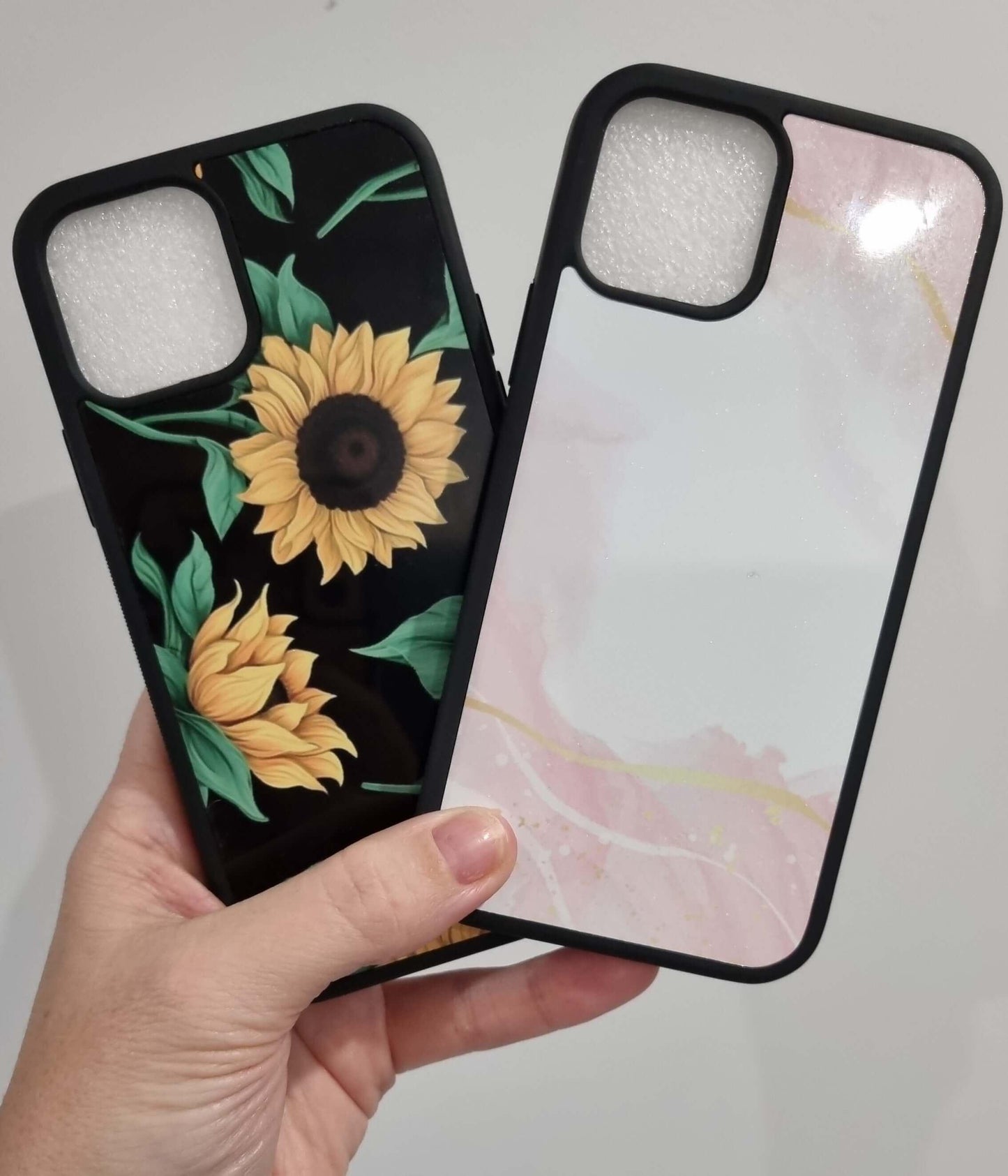 Hand holding two iPhone 15 shock-proof custom design cases, one with sunflowers and one with a pink marble pattern.