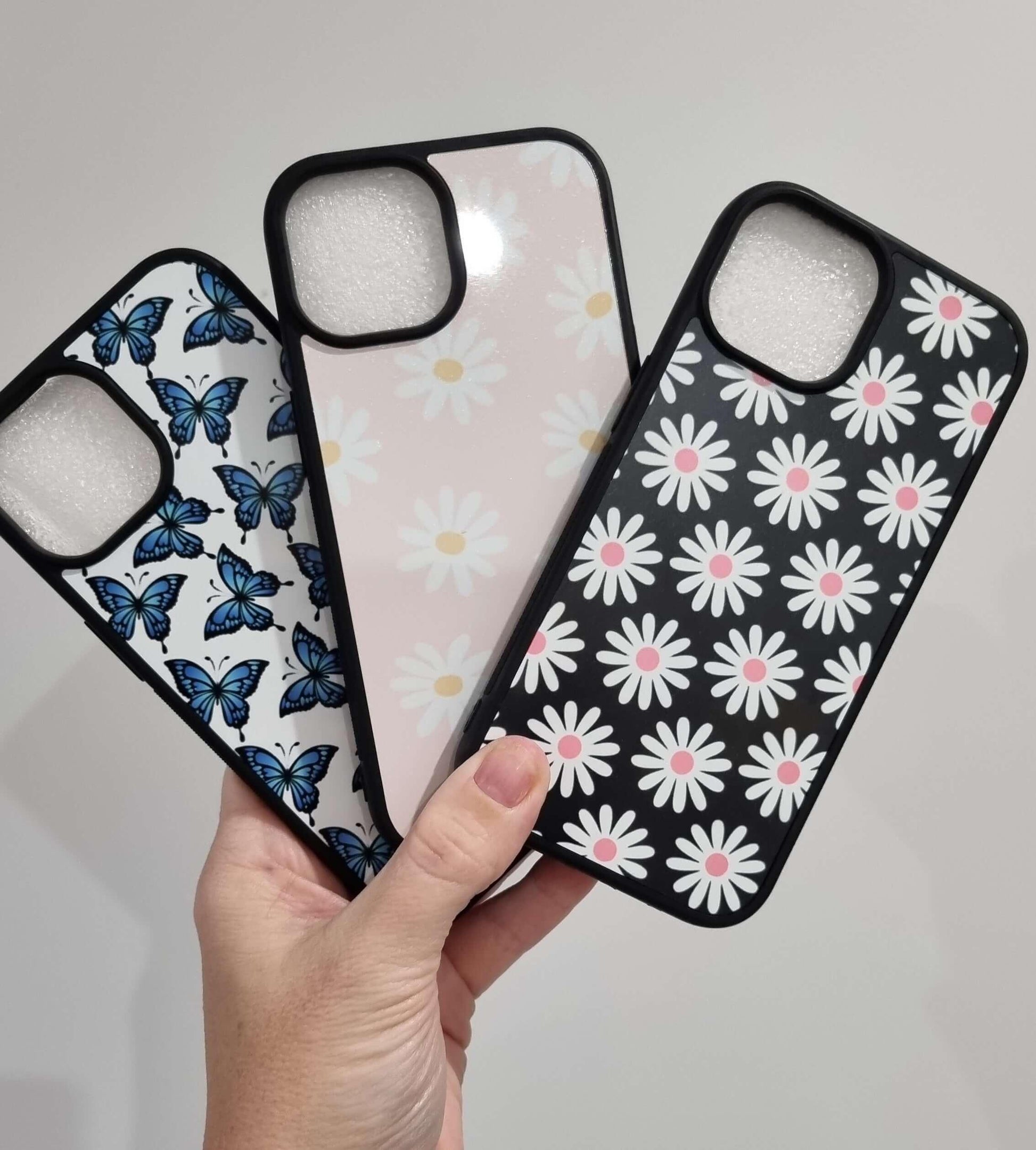 Hand holding three iPhone 15 cases with custom shock proof designs featuring butterflies and daisies.